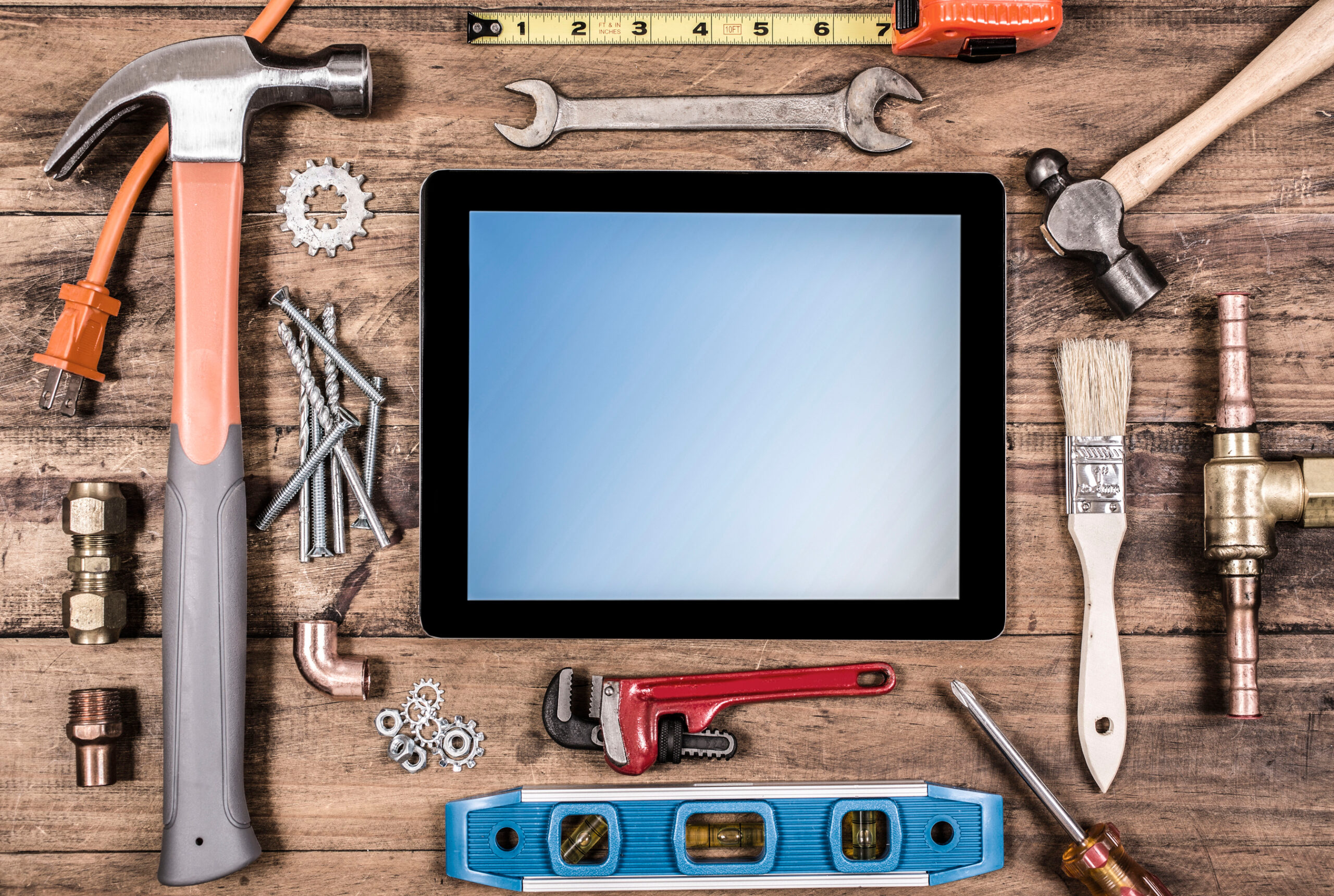 Various construction, DIY hand tools surround a digital tablet.  The screen is blue, blank on the tablet.  Tools include: hammer, nails, level, tape measure, paint brush, wrench, screwdriver.  All items lie on top of a rustic wooden table or desk.  Home improvement, construction themes.  Great background.
